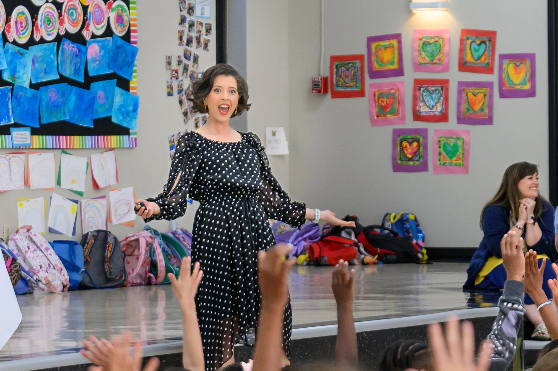 Lisette on stage in the Lower School, smiling toward a row of children whose hands reach up from below; student artwork covers the wall behind her.